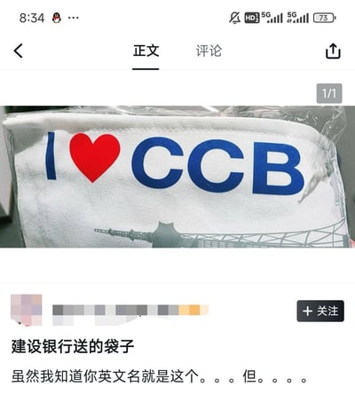 社交媒体帖子截图，内容为一张白色袋子的照片。袋子上印有蓝色的‘CCB’字母和红色爱心图案，下方有模糊的地标建筑线条。图片下方配文：‘建设银行送的袋子 虽然我知道你英文名就是这个。。。但。。。。’，暗示因‘I ❤️ CCB’的设计产生的幽默或略带尴尬的感觉。
