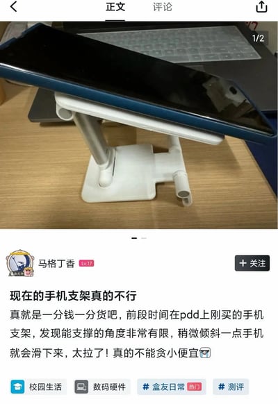 这是一张竖屏的社区帖子截图，上半部分为实拍图：木质桌面上放置着一个白色可升降调节的手机支架，一台带蓝色保护边框的熄屏手机从支架上滑落，刚好卡在支架下方的开口缝隙中，背景还能看到一台带键盘膜的笔记本电脑；下半部分是帖子正文内容，发帖用户ID为马格丁香，等级为Lv17，正文吐槽自己前段时间在拼多多贪便宜购买了这款手机支架，称该支架可支撑的角度非常有限，手机稍微倾斜就会滑下来，感叹一分钱一分货，不该贪小便宜，帖子下方带有校园生活、数码硬件、盒友日常、测评四个话题标签。