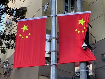 图片呈现了一处城市居民区的户外场景，两根金属杆上悬挂着两面五星红旗。左侧的红旗平整舒展，五星图案清晰规范；右侧的红旗出现明显褶皱，一个白色的监控摄像头恰好卡在红旗的褶皱部位，视觉上仿佛被红旗半包裹住。背景是米黄色的居民楼外墙，墙面上可见空调外机、交错的电线等设施，充满了生活化的日常氛围。