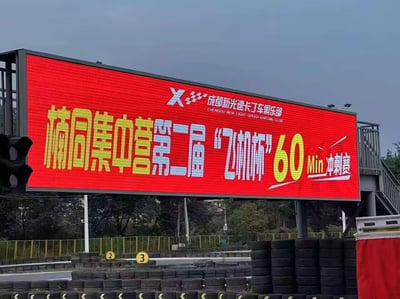 这是一张在成都新光速卡丁车俱乐部户外赛道拍摄的照片，画面主体是赛道上方的大型红色电子显示屏，屏幕上用黄、蓝、白配色的醒目字体展示着赛事信息：‘楠同集中营第二届“飞机杯”60Min冲刺赛’，屏幕右上角标注了俱乐部的中英文名称。屏幕下方是卡丁车赛道的轮胎护栏，背景能看到树木和赛道设施。
特别说明：赛事名称中的‘飞机杯’使用了谐音梗，与成人用品同名，让严肃的卡丁车赛事名称变得趣味搞笑，充满玩梗的氛围。