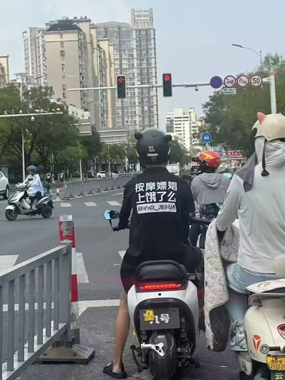图片拍摄于城市路口红灯等待时，画面中最显眼的是一位骑着电动摩托的男性，身穿黑色短袖，背后大字写着“按摩嫖娼上瘾了么，救心点一推时公”，让人发笑又无语。旁边还有其他骑电动车的人，有人穿着动物造型头盔和带有动漫风格的被子，整体氛围轻松搞怪。短袖上的文案本身带有谐音梗和网络用语，内容出其不意，典型街头幽默。