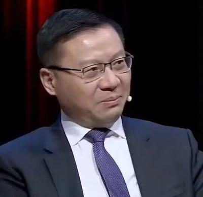 图中人物为张维为，他身着深色西装、白色衬衫，搭配紫色暗纹领带，佩戴黑框眼镜，嘴边夹着节目录制用的领麦，侧脸朝向画面右侧，脸上露出略带玩味、不屑的似笑非笑表情，背景为暗色调，左侧可见部分红色幕布，是传播度很高的张维为梗图素材。