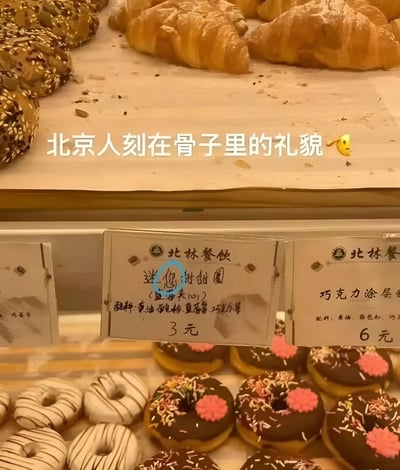 图片展示了面包店的陈列柜，上层摆放着金黄色的羊角面包，下层是装饰精美的甜甜圈（包括白色条纹和巧克力涂层带粉色花朵样式）。陈列柜上的价格标签写有“迷你甜甜圈（望海来口） 3元”和“巧克力涂层 6元”，标签上方有“北林餐饮”字样。图片上方叠加文字“北京人刻在骨子里的礼貌🤔”，幽默调侃标签上“望海来口”的亲切表述，体现北京式的礼貌与热情。