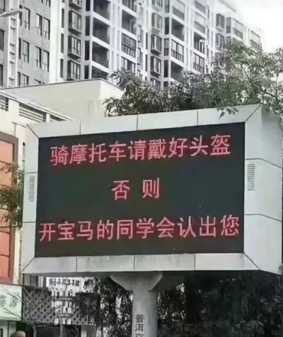 这是一张户外原始拍摄的静态照片，画面主体是一个立在城市户外的电子宣传显示屏，屏幕上用醒目的红色字体分三行展示宣传内容：“骑摩托车请戴好头盔”“否则”“开宝马的同学会认出您”。该标语没有采用严肃的说教式内容，而是用一种带有调侃、接地气的幽默方式，以“怕被开豪车的老同学认出”的趣味角度，提醒骑摩托车的市民佩戴头盔，既贴合生活场景，又能让人印象深刻，达到交通安全宣传的目的。显示屏背景是高层居民住宅楼和一些绿植，显示屏下方的立柱上印有“普洱”字样，表明拍摄地点大概率是云南普洱地区。