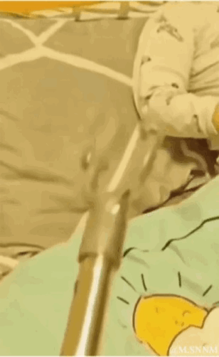 这是一个视觉错位的动态GIF，画面里一根金属杆子的位置和后方坐在床上的人形成视觉错觉，看起来像是杆子直接戳进了人的身体里，实际只是拍摄角度造成的错位效果，制造出荒诞搞笑的视觉反差，没有双关或谐音梗的设计。