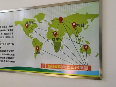 这是胜和餐饮集团的企业宣传展示板，以世界地图为背景，用定位标注出集团业务覆盖的黑龙江、吉林、辽宁、内蒙古四个地区，并用连线展示区域间的关联；左侧配有文字介绍企业发展与业务，底部标注“胜和餐饮|集团现已覆盖”的品牌标语，整体用于展示企业的业务布局与规模，风格正式专业，属于企业宣传物料。