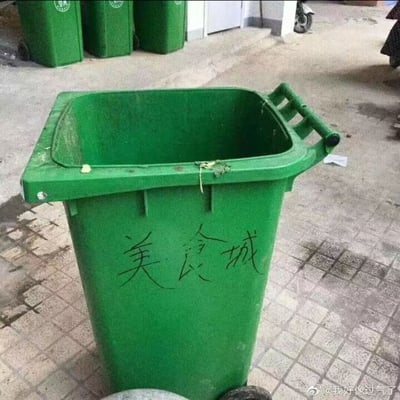 这是一张室外场景的实拍照片，主体是一个绿色的大型户外垃圾桶，桶身外侧被人用黑色笔迹手写了‘美食城’三个字，垃圾桶的边缘还残留着一些食物残渣，呼应了‘美食城’的调侃。背景中可以看到另外几个同款绿色垃圾桶，地面是砖石材质的室外路面，整体场景是日常的户外公共区域。这张图用反讽的方式，将原本用来盛放美食城剩余食物垃圾的垃圾桶直接标注为‘美食城’，制造出强烈的反差感，带有戏谑、调侃的意味，吐槽美食城的食物最终都沦为垃圾的现实。