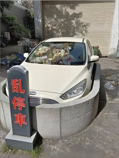 一辆白色福特小轿车被停放在一个圆形混凝土围栏内，车头正前方立着一块灰色石碑，碑上用红色大字写着“乱停车”。汽车的挡风玻璃上堆满了各种杂物和疑似垃圾的物品，周围地面散落着一些废弃物，背景是一扇关闭的卷帘门和几株绿植。整个场景通过“乱停车”标识与被垃圾包围的汽车形成幽默对比，仿佛是对违规停车行为的一种讽刺性“惩罚”。