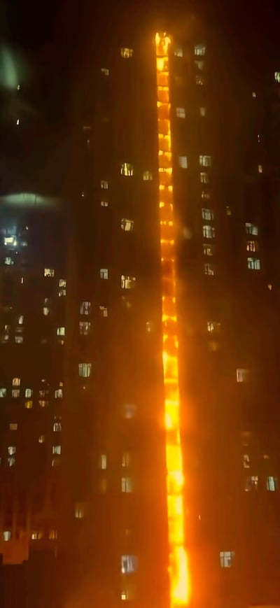 这是一张夜晚拍摄的高层居民楼照片，楼体中间出现一道贯穿上下的明亮火光，呈现出垂直蔓延的态势，推测是楼体外墙相关材料引发的火情。周围楼体的窗户有不少亮着灯光，在漆黑的夜色中，这道显眼的火光凸显出火情的危急，也让整个场景充满紧张感。
