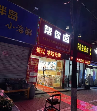 夜晚的城市街道场景，多家店铺招牌亮起霓虹灯。左侧是蓝色招牌的“半岛小海鲜”，中间为红色招牌的“帮你卤”卤味店，门口挂着“错过 欢迎光临”的黄色灯箱，店内可见卤味陈列，旁边贴有“盛大开业 钜惠来袭”的红色海报。右侧有“养生磨坊”等店铺，部分门面卷帘门关闭，贴有“门面出租”字样。整体呈现出热闹的市井夜间氛围。