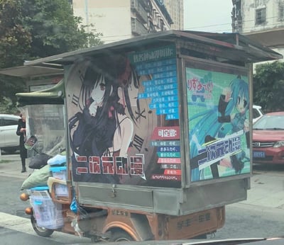 这是一张在城市街头拍摄的照片，主体是一辆经过改装的三轮移动售卖车，被打造成流动的二次元动漫周边摊位。车身外侧张贴着大幅动漫主题广告，左侧是《约会大作战》中的角色时崎狂三，背景是时钟图案，下方印有“二次元动漫”字样；右侧是虚拟歌手初音未来的形象，同样标注“二次元动漫”。车身侧面还详细列出了售卖的各类动漫周边商品，包括徽章、卡贴、手办、古风发饰、围巾、手套等，同时提供定制服务，可定制卡贴、抱枕、不同类型的马克杯、T恤、卫衣等，并附带了联系QQ号。售卖车底部印有“宇丰车业”的字样，车旁堆放着一些货物，背景是城市街道，能看到其他车辆和行人，整体呈现出一个面向动漫爱好者的流动街头摊位的场景。