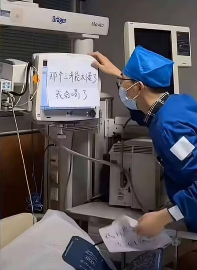 整张图的场景为医院诊疗室，可见多台Drager品牌的专业医疗设备，一名身着蓝色医护工作服、佩戴蓝色手术帽、口罩和眼镜的男性医护人员，正将一张手写白纸贴在医疗监护仪屏幕上，白纸上手写文字为“那个三升袋太慢了 我给喝了”。三升袋是临床用于给患者静脉输注的大容量输液制剂，不可能直接饮用，该内容属于医护人员的趣味玩梗，用不符合常识的表述制造出强烈的反差笑点，十分诙谐。