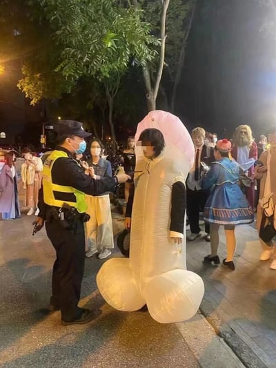 夜晚的街道上，一名穿着反光背心的警察正在与一名身着巨大白色充气服装的人互动。周围有许多穿着各式服装的人，显然是在参加cosplay活动或万圣节庆祝。警察似乎在用手持设备检查或记录什么，而穿充气服装的人则平静配合。背景中可以看到树木和其他围观的行人，整个场景充满了节日的欢乐氛围和一丝滑稽感。
