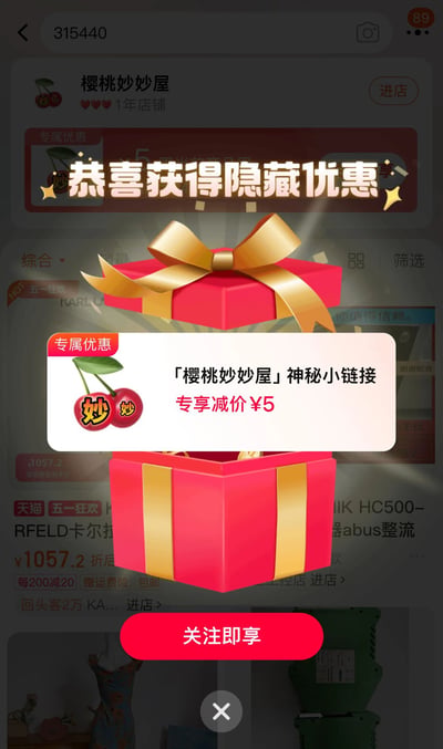 这是一张手机购物APP的弹窗截图，展示了「樱桃妙妙屋」店铺的隐藏优惠活动。画面中央是一个红色礼盒，带有金色蝴蝶结，礼盒上方有「恭喜获得隐藏优惠」的金色文字。弹窗内容显示「『樱桃妙妙屋』神秘小链接 专享减价¥5」，底部有一个红色的「关注即享」按钮。背景隐约可见店铺页面，显示这是一家1年店铺，有「天猫 五一狂欢」等促销信息。