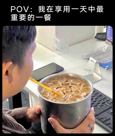 这是一张职场场景的搞笑梗图，画面中一位职场人坐在办公桌前，双手捧着一个大号不锈钢容器，容器里装满了加大量冰块的冰咖啡，正用吸管饮用。图片上方配文“POV：我在享用一天中最重要的一餐”，用夸张戏谑的方式调侃当代打工人对咖啡的重度依赖，将咖啡视为支撑日常工作的“最重要一餐”，办公桌上的键盘、手机、文件架等物品也还原了真实的办公室环境，精准戳中职场人的日常状态，容易引发职场群体的共鸣。