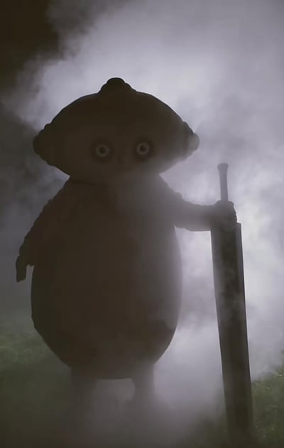 这是一张对童年IP《花园宝宝》角色玛卡巴卡的二次创作梗图，原本软萌可爱的玛卡巴卡被置于昏暗的烟雾场景中，一只手握着一把大剑，昏暗的光线与弥漫的烟雾烘托出严肃硬核的氛围，和角色原本的低幼可爱形象形成强烈反差，制造出荒诞搞笑的效果。