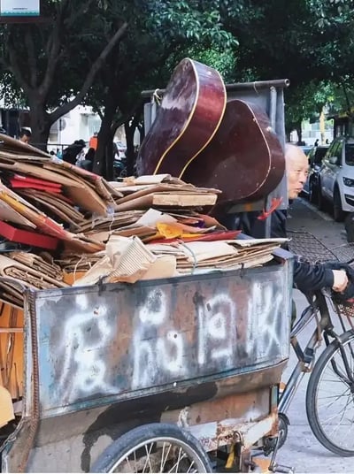 这张街拍照片展示了一辆收废品的三轮车，车上堆满了纸板以及两把吉他盒，形成强烈对比。吉他本是音乐、文艺的代表，而在此则和废品纸盒一样被随意堆放，传递出现实与理想的碰撞感。一边是辛苦拉车的拾荒者，一边是沦为废品的吉他，画面有些荒诞又带些尴尬的幽默。

若细品，有暗示“梦想和生活”的反差感，是一种都市文艺与现实压力的双关隐喻，充满都市生活的心酸与无奈。