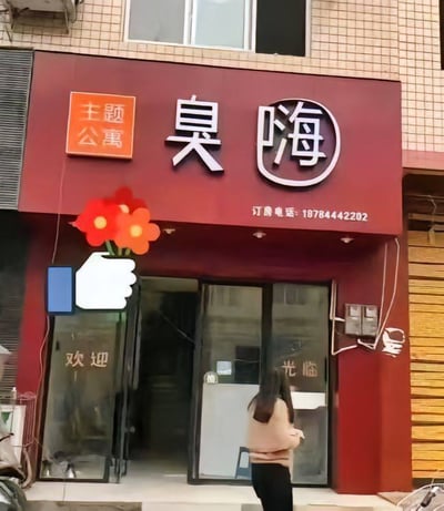 这是一张实地拍摄的街边店铺照片，主体是一家名为‘臭嗨’的主题公寓店面。深红色的店铺招牌十分醒目，左侧橙色方块内标注‘主题公寓’，中间是白色大字店名‘臭嗨’，其中‘嗨’字被圆形边框圈起，招牌下方印有订房电话18784442202。店铺的玻璃门内侧分别贴有‘欢迎’‘光临’字样，门前有一位长发女子正走向店铺入口。此外，图片上还添加了后期贴纸：一个卡通手举两朵红花的图案，以及一个蓝色方块搭配白色点赞手势的图案。店名‘臭嗨’运用了谐音梗，在部分方言中‘臭嗨’是脏话，此处用作店名，大概率是为了制造反差感和趣味性，吸引路人注意。