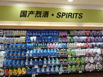 这是一张超市内部的实拍图，货架顶部的浅绿色背景标识牌上用黑色字体标注着“国产烈酒 · SPIRITS”，但标识牌下方的多层货架上整整齐齐摆放的全是不同品牌、不同规格、不同颜色包装的洗衣液、洗衣凝珠等衣物清洁洗护产品，标识标注的商品品类和实际售卖的商品完全错位，产生了滑稽的反差效果。