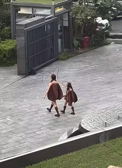 图中展示了两个穿着棕色蚂蚁服装的人——看起来像一位成人和一个小孩——正背对着镜头向前走。她们的服装带有长长的黑色触角，下身搭配深色靴子。背景是一个带有黑色铁门的入口，周围有茂密的绿植和停放的车辆，地面是灰色的石板铺成，整体场景像是在某个小区或别墅区的户外区域。