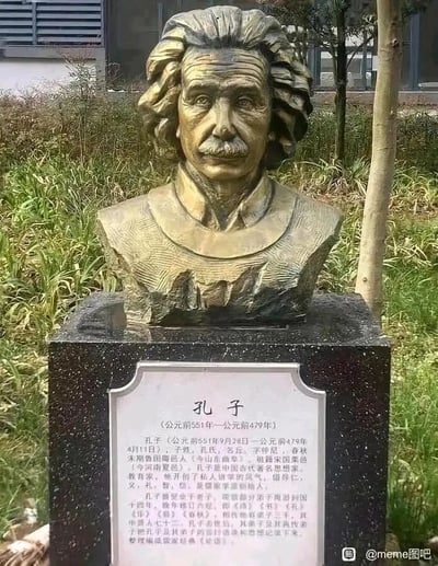 这张图片呈现了一个荒诞滑稽的户外场景：一尊外观为著名物理学家阿尔伯特·爱因斯坦的半身青铜雕像，有着他标志性的蓬松卷发和胡须，面部表情严肃，以静止的姿态立在黑色石质基座上。但基座正面的白色铭牌上，却详细印着中国古代著名思想家、教育家孔子的生平介绍，包括孔子的生卒年月、字号、籍贯、历史地位、学术贡献、相关著作等完整内容，将两个毫无关联的中外名人的形象与介绍错误配对，形成强烈的反差感，制造出荒诞搞笑的效果，属于典型的张冠李戴式梗图。
