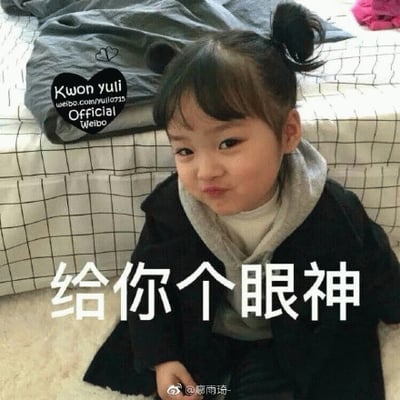 图片中是韩国童星权律二，她扎着两个小巧的丸子头，身穿黑色外套内搭灰色连帽衫，坐在铺有白色地毯的地面上。背景可见带有网格图案的床铺和灰色被子，她歪着头，嘴角微微上扬，眼神灵动，做出俏皮搞怪的表情，下方配有醒目文字“给你个眼神”，整体画面充满童真与趣味性，展现了其标志性的萌系风格。