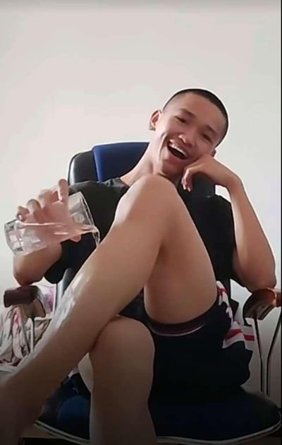 图片中一名男子坐在蓝色办公椅上，双腿交叠，姿态十分放松。他穿着黑色T恤和深色短裤，左手撑着脸，右手拿着一个透明玻璃杯，杯中装有少量液体。男子面带夸张的笑容，嘴巴大张，眼睛微眯，看起来非常开心。背景是简单的白色墙面，整体氛围轻松搞笑，适合作为表达开心、得意情绪的表情包使用。