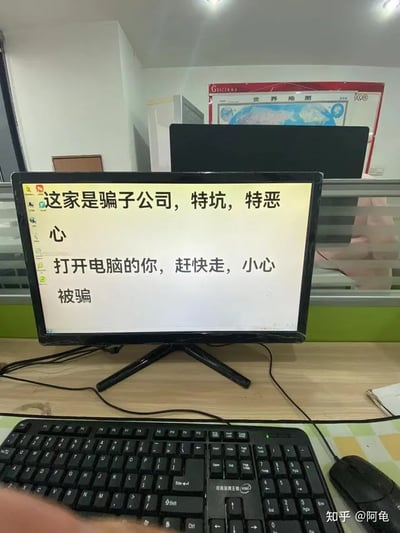 这是一张办公室场景的实拍图片，画面中心是一台放置在办公桌上的电脑显示器，屏幕上显示着一段醒目的警示文字：“这家是骗子公司，特坑，特恶心 打开电脑的你，赶快走，小心被骗”，推测是遭遇了不良职场待遇的前离职员工留下的提示，目的是提醒后续使用该电脑的新入职人员或访客，警惕这家公司的欺骗行为。显示器下方配有黑色的键盘和鼠标，画面左下角能看到一小部分人的手部，背景中可见办公室的玻璃隔断、张贴的世界地图以及其他办公设备，图片右下角带有“知乎@阿龟”的水印，表明了图片的来源。
