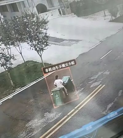 图片展示了一个道路场景，背景中有树木和建筑物。画面中央偏左位置有一个红色边框的标语牌，上面写着“非机动车不戴安全帽”。标语牌下方有一个橙色边框的特写 inset，显示一名身穿白色上衣的人正坐在一个绿色的垃圾桶上。主画面可能是交通监控或街头摄像头拍摄的截图，底部有一条蓝色横幅，上面有模糊的白色文字。整体场景似乎在通过特写 inset 与标语形成对比，突出某种不规范行为。