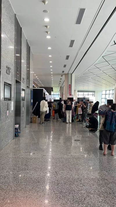 这是一张公共场所走廊的实拍图，画面中可见一条长长的走廊，左侧是灰色大理石墙面，墙上有编号为“2-1”等的电梯门，电梯旁有小型显示屏。走廊地面为光滑的水磨石材质，反射着天花板的灯光。大量人群在走廊中间排队，有人站立等待，有人手持行李，部分人蹲坐在地上，整体场景呈现出忙碌的等待氛围。
