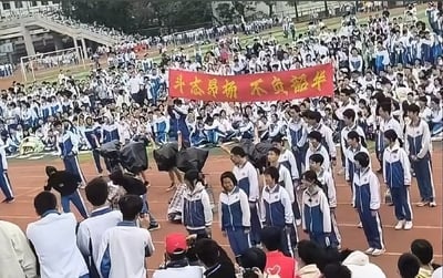 图片展示了一所学校的大型集体活动场景，操场上聚集了大量身着蓝白相间校服的学生，人群密集且有序。画面中央有几名学生共同举着一条红色横幅，上面用黄色字体写着“斗志昂扬 不负韶华”的标语。部分学生弯腰似乎在整理物品或进行准备工作，整体氛围热烈而庄重，推测可能是学校运动会、开学典礼或其他大型集会活动。