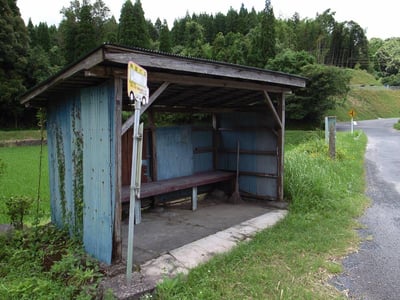 这是一张实拍的日本乡村场景图：画面主体是一个充满岁月痕迹的老旧公交站台，以木质框架搭建，搭配褪色泛旧的蓝色波纹金属板围挡，站台内设有深色长凳，角落还放置着一把扫帚，围挡侧壁爬着藤蔓，尽显质朴的年代感。站台旁立着带有日文标识的「林田川2」站牌，一侧是绿意盎然的稻田，另一侧是蜿蜒的乡村公路，远处是茂密的山林，整体氛围宁静治愈，完美展现了日本乡间的恬淡日常。