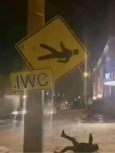 这是一张夜晚户外街道的实拍图，画面左侧的路杆上，下方是一块印有“IWC”字样的黄色标识牌，上方是一块黄色的行人摔倒警示标志，标志上的黑色人形呈向前摔倒的姿态；而在画面下方的地面上，恰好有一个真人以和警示标志上几乎完全一致的姿势躺倒在地，形成了极具巧合感的荒诞画面。同时“IWC”字样很容易让人联想到“WC（厕所）”，结合摔倒的姿势，仿佛是警示标志里的人形“掉”到了现实中，进一步放大了画面的搞笑效果，让整个场景充满了无厘头的趣味。