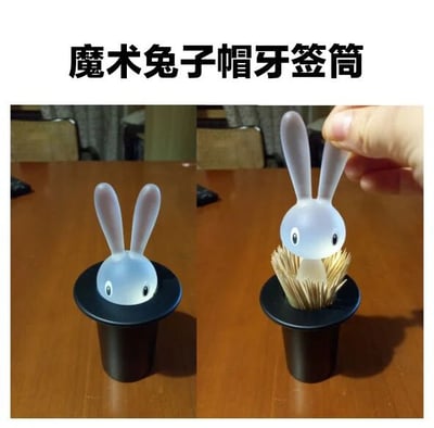 这是一张左右对比的产品展示图，展示了一款名为“魔术兔子帽牙签筒”的创意家居用品。左侧图片中，一个黑色圆柱形的“魔术帽”容器上，有一个白色半透明的兔子头造型部件安放在顶部，兔子眼睛为黑色圆点，显得呆萌可爱。右侧图片中，一只手正捏住兔子头将其向上提起，此时容器内部露出整齐排列的牙签，直观展示了其作为牙签筒的实用功能。图片上方配有产品名称文字“魔术兔子帽牙签筒”，整体构图清晰，突出产品的设计巧思和使用方式。