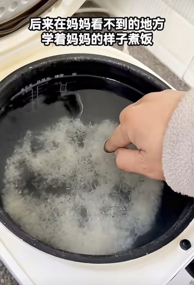 这是一张生活实拍图，画面展示了打开的电饭煲内胆，其中盛放着正在淘洗的大米和清水，一只穿着浅灰色毛绒袖套的手正将手指插入米水中，模仿长辈常用的手指测量水位的方法准备煮饭，图片顶部配有文字“后来在妈妈看不到的地方学着妈妈的样子煮饭”，充满生活感，能够引发独自生活的年轻人的情感共鸣。