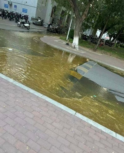 这是一张户外实拍的城市道路场景图，画面核心区域的路面出现塌陷，形成了一处深坑，坑体及周边路面被浑浊泛黄的积水覆盖，积水几乎淹没了深坑的边缘，使得这处路况隐患变得十分隐蔽。画面左侧是红砖铺设的人行道，人行道旁的树木底部被涂成白色；画面远处可见一栋建筑物，建筑外停放着多辆电动车、摩托车，还有一辆小型拖车，整体场景大概率位于城市的居民区或街巷区域，这样的状况会给过往行人和车辆带来极大的安全风险，容易引发意外。