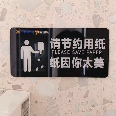 这张照片拍摄的是安装在水磨石墙面上的一块黑底白字节约用纸提示牌，提示牌左上角印有招商蛇口和招商花园城的品牌标识，左侧配有简笔小人抽取纸巾、碎纸掉落的示意图标，右侧上方是“请节约用纸”的中文提示语及对应的英文翻译“PLEASE SAVE PAPER”，下方则使用了谐音梗文案“纸因你太美”，该文案谐音自网络热门梗“鸡你太美”，用趣味化的方式呼吁公众节约用纸，图片左下角还露出了部分其他公共设备的边缘，左下角有标注“Vbitbh.”的小字水印。