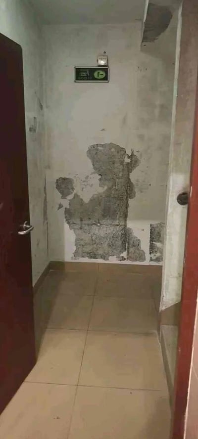 这是一张在狭窄楼道内拍摄的照片，楼道地面铺着米黄色地砖，左侧是一扇打开的深色木门，前方的墙面出现大面积墙皮脱落，露出的水泥基层与残留墙皮恰好组成了类似奥特曼的站立形象，墙面上方还安装着一块绿色的安全出口指示牌（带有向下箭头与“安全出口”字样）。整体场景显得较为老旧，而墙皮脱落形成的巧合图案让普通的楼道场景变得充满趣味，属于偶然发现的生活趣味瞬间。