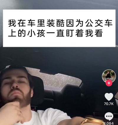 这是一张来自短视频平台的视频截图，画面主体是一名留着胡须的男子，他坐在汽车内，单手托着下巴，摆出严肃耍酷的表情。画面上方配有中文文字“我在车里装酷因为公交车上的小孩一直盯着我看”，解释了他装酷的原因，这种成年人因孩童注视就刻意摆酷的行为充满了反差感，显得幼稚又搞笑。画面右侧是短视频平台的UI元素，显示该视频获得了70.7K的点赞，1084条评论，还有用户的头像及关注按钮，表明这是一个面向大众的趣味日常分享视频。