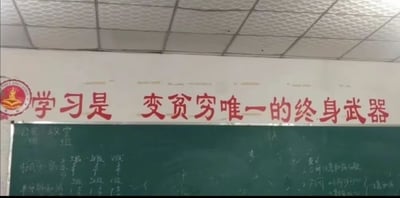 这是一张教室场景的实拍照片，画面上方是方格吊顶，下方黑色黑板上方的白色墙面上印着红色大字标语，内容为「学习是 变贫穷唯一的终身武器」，标语左侧还有一个学校的圆形校徽，黑板上留有未擦干净的模糊粉笔板书，标语内容和传统鼓励学习脱贫的标语反向，充满扎心反讽的效果。