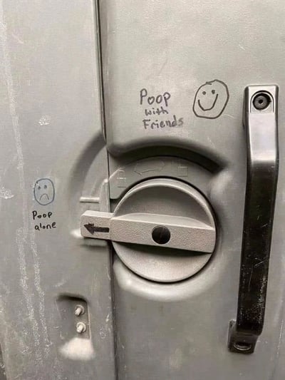 The image shows a gray metal bathroom stall door with handwritten graffiti. On the left side, there's a sad face drawing with the text 'Poop alone' below it. On the right side, a smiley face drawing is accompanied by the text 'Poop with Friends' above it. The center of the door features a circular lock mechanism with a lever and an arrow pointing to the left. The graffiti humorously contrasts the experience of using the bathroom alone (sad) versus with friends (happy).