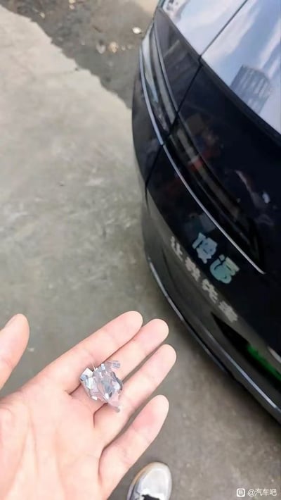 一只手握着一块破碎的锋利玻璃碎片，背景是一辆黑色汽车的后部。汽车车身上用绿色字体写着‘傻运 让我先富’的字样，地面为灰色水泥地，带有一些污渍。图片可能意在通过汽车上的文字与碎玻璃形成对比，营造一种讽刺或幽默的效果，暗示‘傻运’带来的可能并非好运，反而导致车窗破碎等意外。