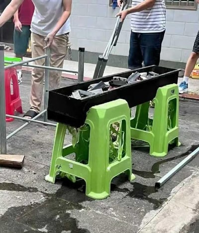 街头场景中，几个人正在搭建一个临时烧烤摊。画面中央是一个黑色长方形烤炉，被架在两个鲜艳的绿色塑料凳上，烤炉内似乎放有木炭或燃料。周围有几个人正在准备，一人手持长柄工具，旁边还有金属支架和红色塑料凳等物品。地面有些潮湿，可能是刚清洗过或雨后。整个场景展现了人们利用简单日常物品即兴搭建烧烤设施的创意。