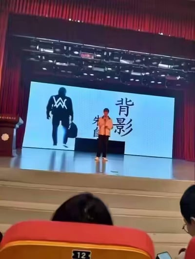 这是一场舞台朗诵活动的现场画面，台上的表演者正在进行朱自清经典散文《背影》的朗诵，但背景大屏上本该是朱自清父亲的背影，被替换成了知名电音制作人Alan Walker的标志性剪影，严肃的文学主题和潮流的电音形象形成强烈反差，制造出搞笑的错位效果，属于二次创作的整活内容。