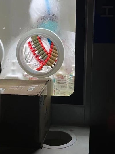 这是一张实验室工作场景的实拍图，画面主体是一个带圆形观察窗的传递窗，窗内放置着多支红帽采血管，被透明塑料袋包裹着；传递窗下方的台面上摆放着两个贴有中文标签的透明试剂瓶，前景处有一个棕色纸箱，台面还有一个圆形孔洞。整个场景呈现出医学检验实验室的日常工作物料存放状态，没有双关、谐音梗或暗示内容。