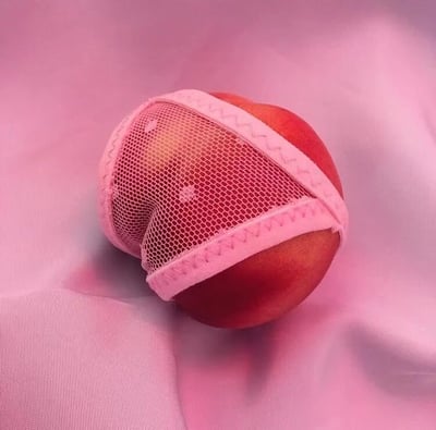 一张创意摄影作品，展示了一个新鲜的水蜜桃被拟人化地穿上了一件粉色网纱小内内。蜜桃呈现出自然的红色渐变，而那件小巧的'内衣'则有精致的蕾丝花边装饰，与蜜桃的形状完美贴合。整个画面以柔和的粉色布料为背景，营造出一种可爱又略带幽默的视觉效果。这种将水果拟人化的创意手法，赋予了普通食材趣味性和话题性。