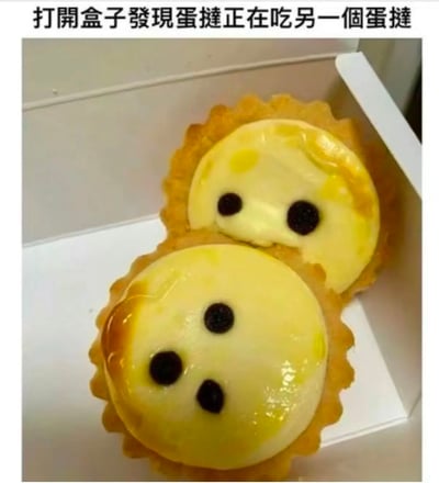 一张创意美食照片，展示了两个放在白色盒子里的蛋挞。上方的蛋挞被摆放在下方蛋挞的顶部，两者都用巧克力豆点缀出眼睛，形成了一个蛋挞正在“吃”另一个蛋挞的有趣视觉效果。图片上方配有文字“打开盒子发现蛋挞正在吃另一个蛋挞”，增强了画面的幽默感和故事性。
