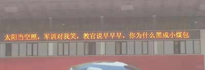 这是一张拍摄校园户外电子显示屏的设备界面截图，属于静态单图，屏幕上用红色字体显示了一段改编自经典儿歌《上学歌》的趣味标语：“太阳当空照，军训对我笑，教官说早早早，你为什么黑成小煤包”。这段文案将军训拟人化，用幽默调侃的语气，精准抓住了学生军训时会被烈日晒黑的普遍经历，把原本平淡的儿歌内容替换成贴合军训场景的内容，充满趣味性，能够缓解军训的枯燥氛围，很容易引发经历过军训的人的共鸣。