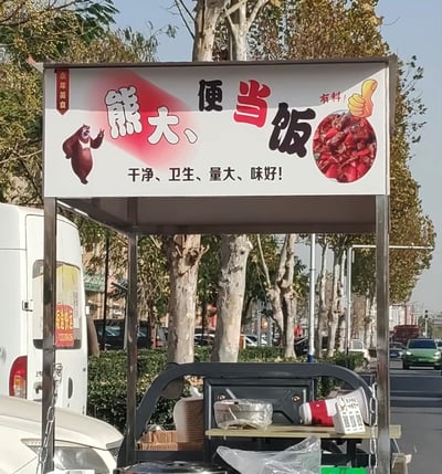 这是一张实拍的街头移动便当摊位的照片，核心内容是摊位上方的白色招牌。招牌左侧印有国产动画《熊出没》中的角色熊大的卡通形象，主视觉文字为“熊大、便当饭”，其中“便当”二字为红色，其余文字为黑色，下方配有宣传语“干净、卫生、量大、味好！”，右上角印有红色餐品的实物圆形配图，旁边标注“有料！”字样和黄色点赞手势图标，左侧还有竖排的“永年美食”红色标识。摊位设置在城市道路旁，背景可见成排的行道树、路边绿植、停靠和行驶的车辆。该招牌的文字排版存在趣味歧义，若忽略文字间的顿号，很容易被误读为“熊大便 当饭”，形成荒诞的搞笑效果。