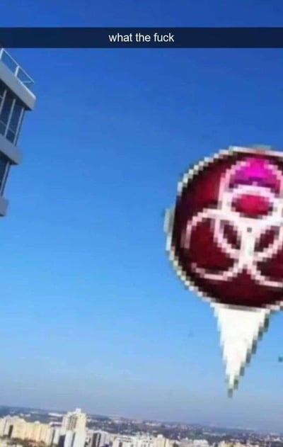 这是一张合成类搞笑梗图，背景是城市高空视角的实拍场景，湛蓝天空下可见成片的城市建筑群。画面右侧被合成了一个像素风格的生物危害（生化危机）标记，标记带有尖刺状底部，仿佛悬浮在城市上空。画面顶部配有白色英文文字“what the fuck”，直白表达出看到这违和一幕的震惊、离谱情绪。
这张图通过将危险题材的图标与日常城市实景强行结合，制造出荒诞戏谑的反差感，没有双关或谐音梗，纯粹靠违和感带来搞笑效果。
